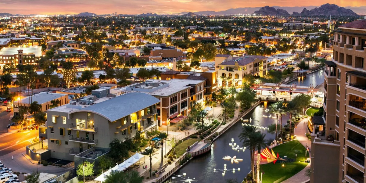 A lively canal scene with modern buildings lining the waterway, twinkling city lights reflecting on the surface, mountains visible in the background, and a striking art installation in the canal. Pedestrians stroll along the waterfront, with outdoor cafes and boutiques nearby, creating an energetic after-dusk atmosphere.