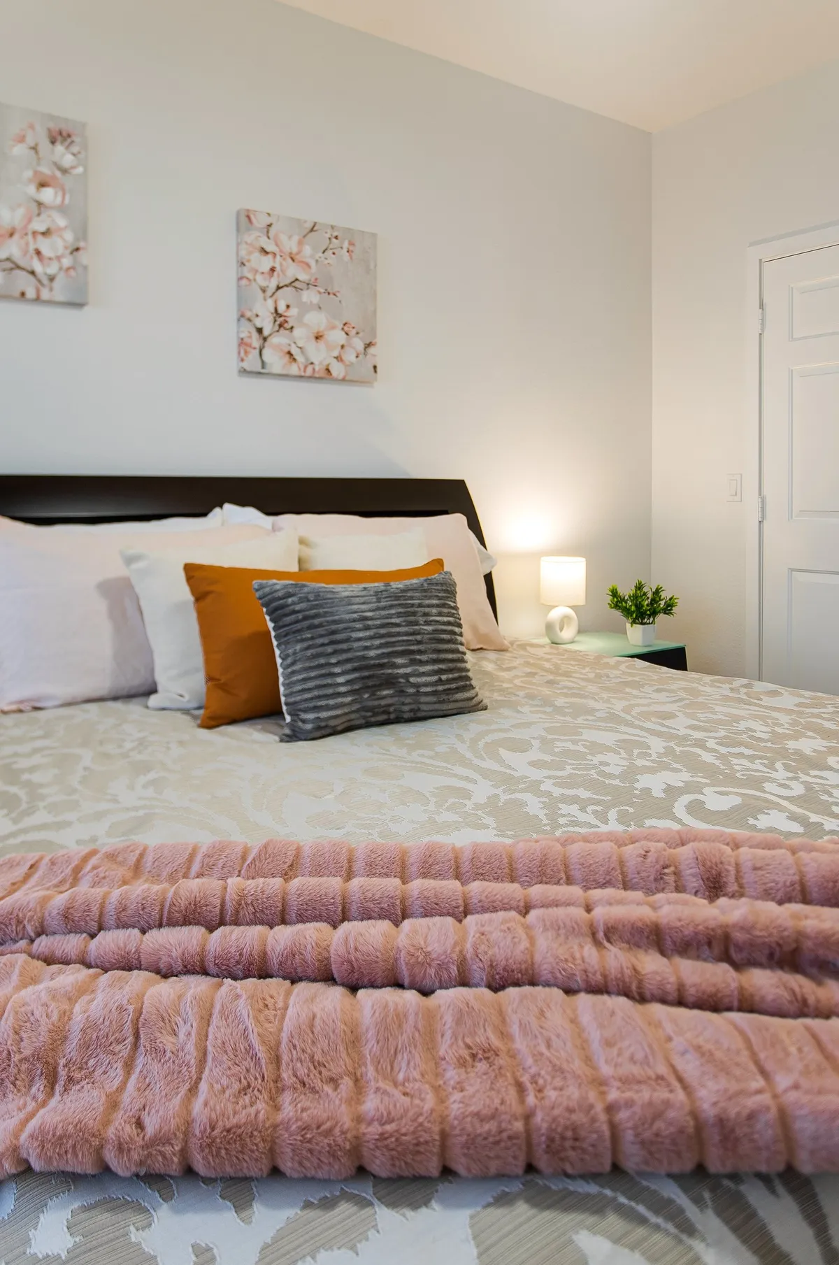 A cozy bedroom with a bed layered in decorative pillows, complemented by floral artwork on the walls and a soft throw blanket at the foot. The ambiance is peaceful and inviting, ideal for curling up with a book or enjoying a quiet moment.