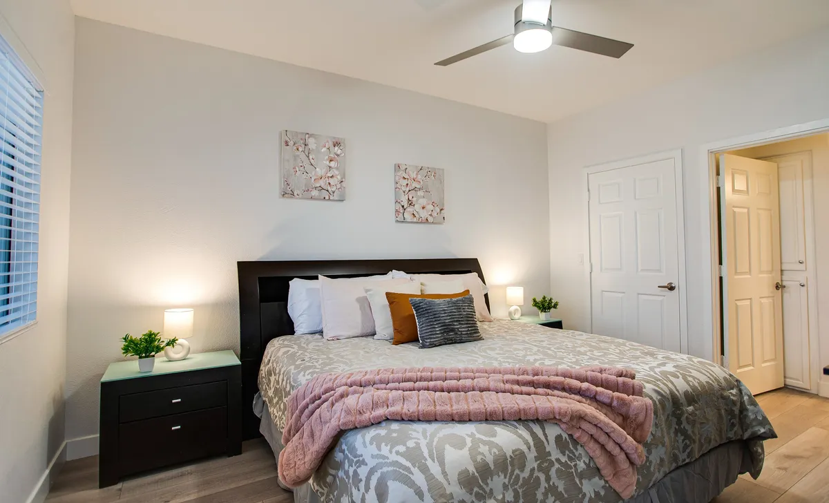 A tranquil bedroom featuring a bed with plush decorative pillows, flanked by nightstands and adorned with calming artwork. Soft natural light streams through a nearby window, creating a cozy atmosphere perfect for relaxation or a restful night’s sleep.