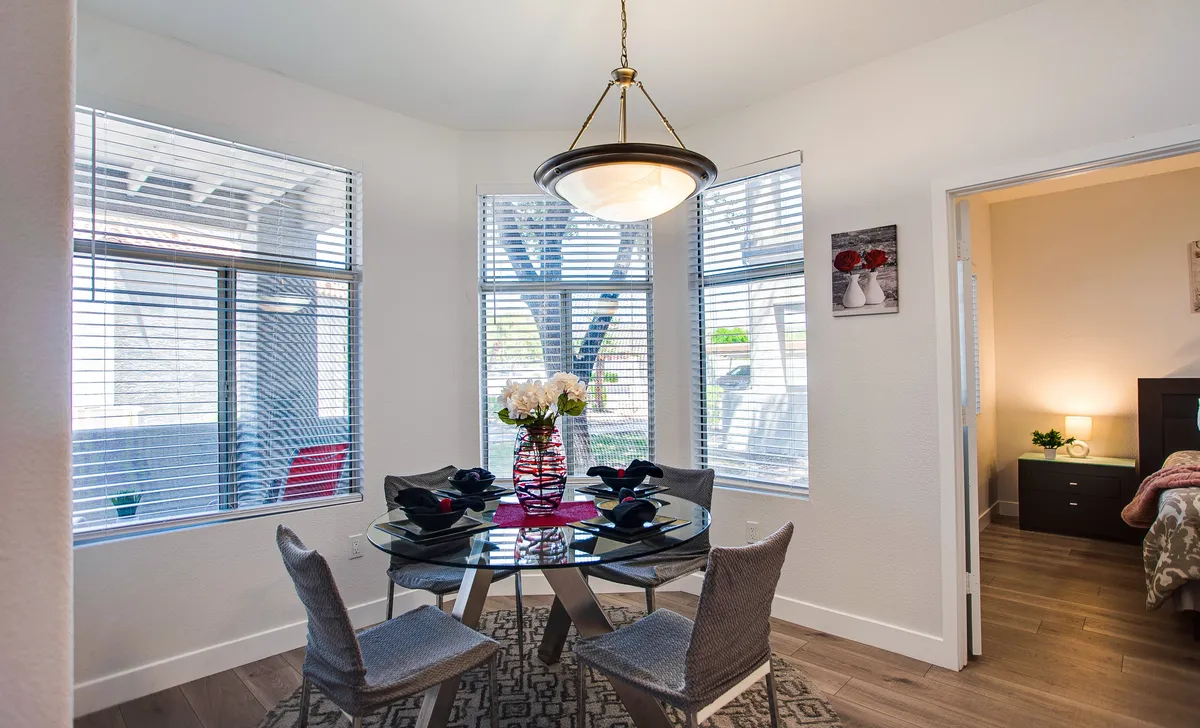 A bright dining area with a round glass table, floral centerpiece, and large windows with blinds. The room feels warm and inviting, ideal for socializing or enjoying a quiet breakfast, and it is filled with natural light.