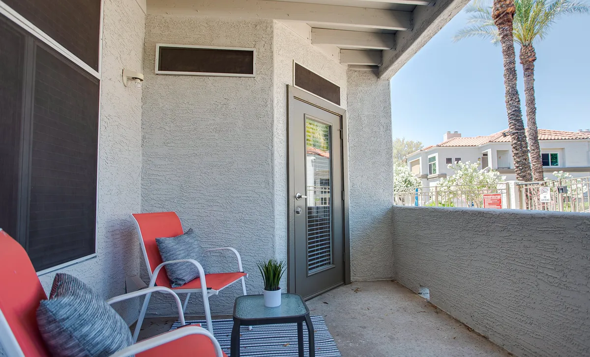 A cozy ground-floor patio space featuring two red chairs, a small table, a potted plant, and a sliding glass door, blending indoor comfort with outdoor ambiance. The setup suggests a tranquil nook for morning coffee or casual conversation, set just steps from the pool area.