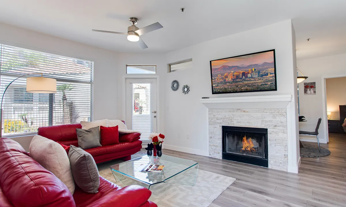 A cozy living area featuring a striking red leather sofa, a glass coffee table, a stone fireplace, and framed cityscape artwork. The setting is modern with warm lighting and invites relaxation or social gatherings.