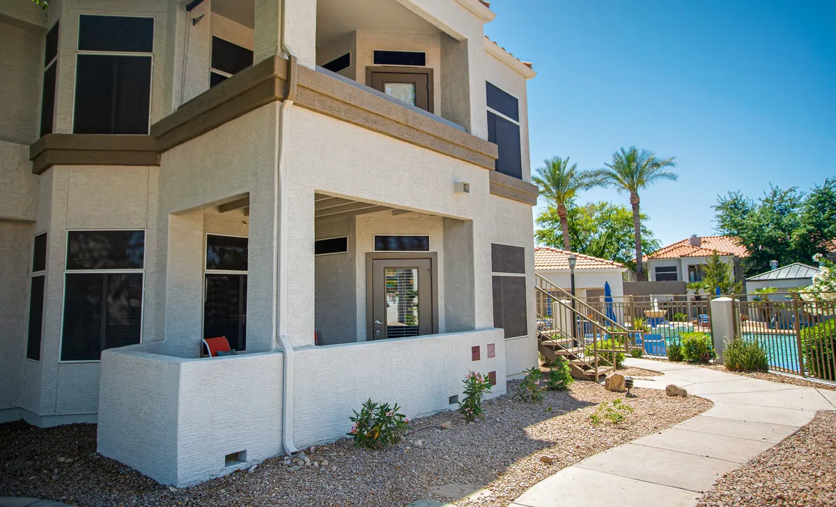 An inviting exterior perspective showing a corner balcony, a picturesque pathway leading toward the pool area, and lush palm trees in the background. The image captures the ease of movement between private and communal spaces, bathed in warm, natural light.